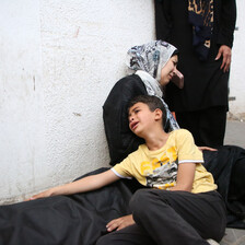 A crying child leans on a woman, sitting next to a corpse.