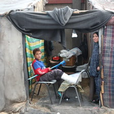 A body with his leg in plastercast sits and a woman stands in a makeshift tent 
