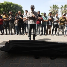 A man stands in front of a dead body in a shroud, with a group of other men behind him