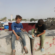 Two boys sit on blocks of concrete with tents behind them 