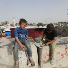 Two boys sit on blocks of concrete with tents behind them