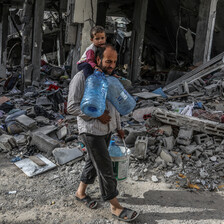 A man carries a child on his shoulders and water containers in his hands as he walks through rubble in southern Gaza