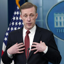 Jake Sullivan holds his hands towards his chest while standing in front of a US flag