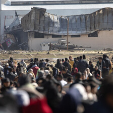 A crowd of people is overlooked by an armored tank in the background