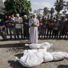 An imam in white leads an outdoor prayer next to two bodies wrapped in white cloth