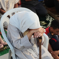 An older person sits, bows their head and leans on a walking stick during prayers in Gaza