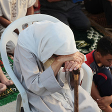 An older person sits, bows their head and leans on a walking stick during prayers in Gaza 
