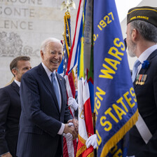 Joe Biden smiles as he shakes hands with a soldier in uniform standing behind a number of flags 