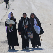 three women walk on a dusty road, one carrying an infant