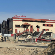 A destroyed wedding hall sits on a dirt road on a bright day with people walking in front