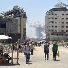 A few people walk beside a badly damaged building with smoke in the background 