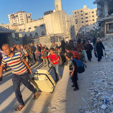 A man in a crowd pulls a suitcase through a neighborhood of destroyed buildings