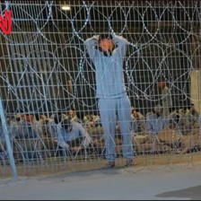 A man wearing a gray tracksuit and with is face mainly covered stands in front of many other men who are seated and behind coils of barbed wire 