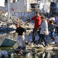 People walk through a pool of sewage water amid the rubble of buildings