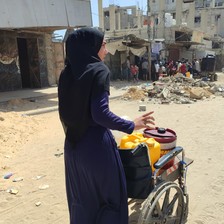 A woman stands behind a wheelchair containing water containers 