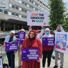 Woman holds sign reading "Protect Gazan healthcare workers" while standing infant of five others holding signs and wearing keffiyehs