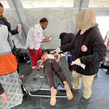 A woman wearing a hoodie with a Red Crescent logo stands over a boy lying face down on a stretcher as other medics work around them