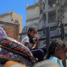 Two girls sit on top of mattresses with destroyed buildings in background