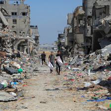 Three men walk in the middle of the road surrounded on either side by destroyed buildings spilling out their household contents