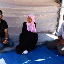 Sitting on a blue cloth under a shade are a young man, a young woman and an older man