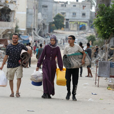 Three people walk with bags and blankets on a backdrop of ruined buildings