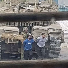 Two men with arms behind their back walk in front of an Israeli tank