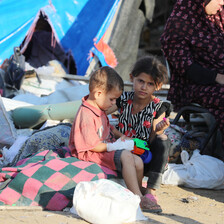 Two children sit in front of a tent that has become their shelter