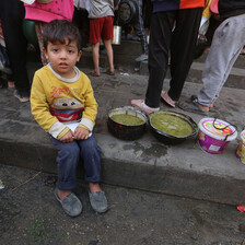 A young Palestinian child sits alongside food as others queue