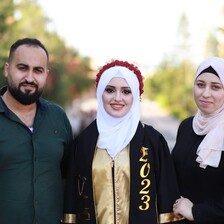 In the daytime, three people smile for the camera, one in graduation gown