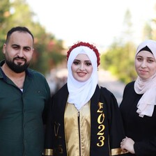 In the daytime, three people smile for the camera, one in graduation gown