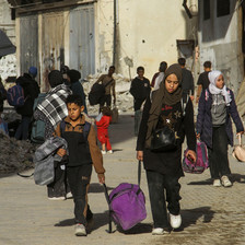 People, including children, carrying their belongings walk down a street strewn with rubble