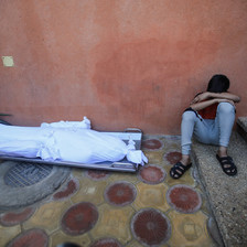 A boy covers his face with his hands while sitting next to three shrouded bodies