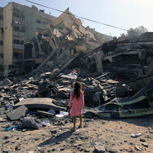 A girl in a pink dress is seen from the back while standing in front of a destroyed building