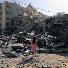 A girl in a pink dress is seen from the back while standing in front of a destroyed building