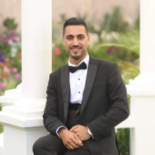 A man wearing a tuxedo poses, sitting and smiling, in a garden.