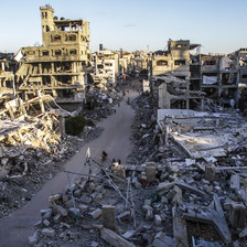 Landscape view of people walking and biking on narrow road cleared of debris between destroyed and bombed-out multi-story buildings