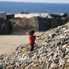 A young girl stands on top of a garbage pile with makeshift shelters and the sea in the background