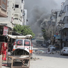 An ambulance in foreground and huge dust cloud in background caused by Israeli bombing