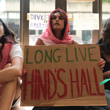 Three young women sit on floor while wearing kaffiyehs and holding sign reading: Long Live Hind's Hall