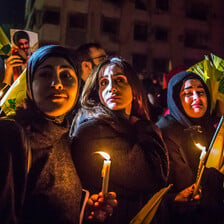 Three young women with sad expressions on their faces hold lit candles and Hizballah flags