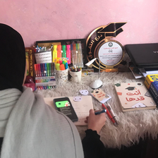 Young woman is seen from back while sitting at desk with a phone, notebook and writing tools