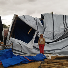 Under cloudy skies, a child stands in front of a collapsed tent