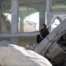 A young woman with a pensive gaze sits on a slab of concrete while holding her chin in her hand