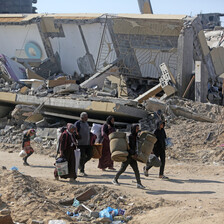 Landscape view of a family of six carrying rolled-up mattresses and buckets while walking on road lined by destroyed buildings