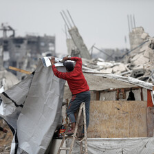 A man nails a tarp cover onto a wooden structure surrounded by rubble