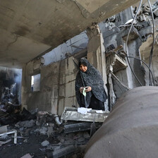 Inside a gutted-out concrete building stands a woman, a plume of smoke and dust rising to her left. 