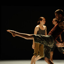 A man lifts his foot as part of a dance routine in front of two other people on a stage