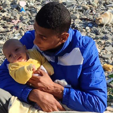 A man in a blue jacket cradles a newborn baby in a yellow jumper amid piles of rubble