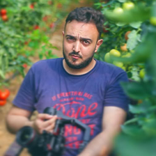 Young man wearing t-shirt holds camera while standing next to tomato plants