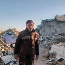 An older man looks at the camera while standing on narrow road in between destroyed buildings